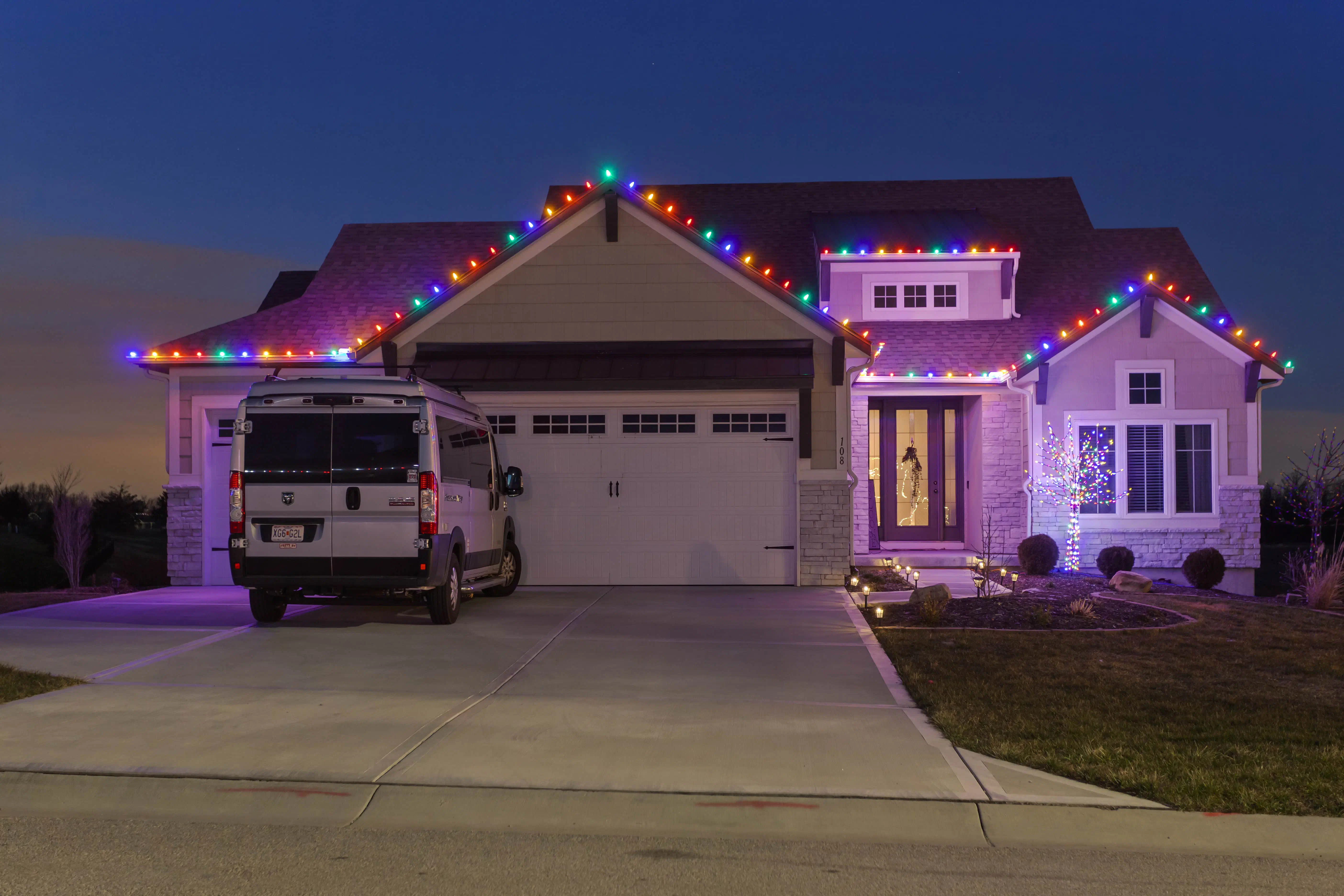 Tis The Season work van parked in front of a home lit with multicolor holiday lights at dusk